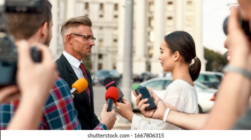 Horizontal Shot Of Serious Politician Giving Interview To Female Reporter. People Making Interview Using Camera And Equipment Set At Outdoor Location. Selective Focus. Side View