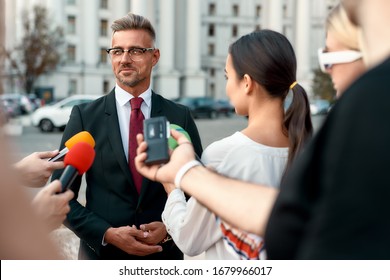 Horizontal Shot Of Serious Politician Looking Away And Giving Interview To Reporters. People Making Interview Using Microphones And Equipment Set At Outdoor Location. Selective Focus. Front View