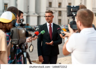 Horizontal Shot Of Serious Politician Looking At Reporters And Giving Interview To Them. People Making Interview Using Microphones And Equipment Set At Outdoor Location. Selective Focus. Front View