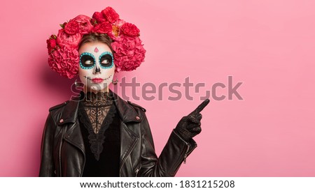 Horizontal shot of serious woman wears scary makeup spends free time on halloween party points away at copy space isolated over pink background celebrates traditional mexical holiday. Face art