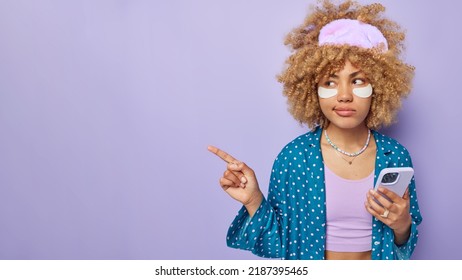 Horizontal Shot Of Serious Young Woman With Curly Hair Points Away On Blank Space Advertises Product Holds Mobile Phone Dressed In Nightwear Isolated Over Purple Background. Look At This Promo