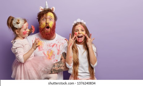 Horizontal Shot Of Shocked Dad Has Yellow Face Painted With Watercolors, Two Children Have Fun With Father, Joyful Expressions, Isolated Over Purple Background With Free Space For Promotion.