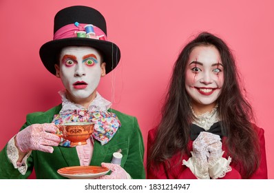 Horizontal Shot Of Shocked Male Hatter Poses With Cup On Tea Party. Happy Brunette Woman Has Spooky Makeup Bloody Wounds On Face. Couple Ready To Go On Halloween Zombie Party. Face Art Concept