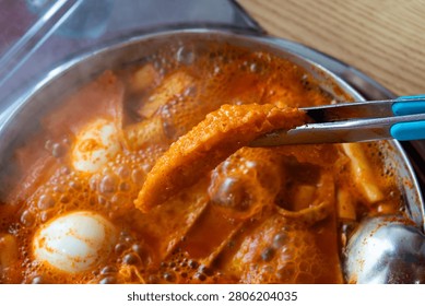 A horizontal shot showing a crispy deep-fried dumpling being lifted by metal tongs over a steaming, boiling pot of Korean spicy rice cakes with visible steam and a transparent lid on the side.