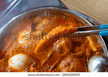 A horizontal shot showing a crispy deep-fried dumpling being lifted by metal tongs over a steaming, boiling pot of Korean spicy rice cakes with visible steam and a transparent lid on the side.