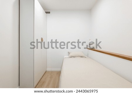 Horizontal shot of a single bed with a spacious white wardrobe, shelf and light fixture against white walls. Concept of an apartment bought with a mortgage for a single person