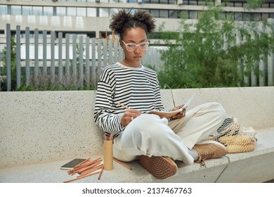 Horizontal shot of skilled creative young woman makes drawings uses colorful crayons wears trendy sunglasses striped jumper and white trousers sits in lotus pose outdoors. People and creativity