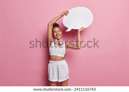 Horizontal shot of slim Latin woman dressed in white sportswear holds blank communication bubble for your advertisement uses stereo headphones isolated over pink background. Sporty lifestyle concept