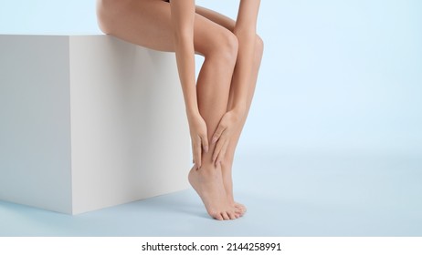 Horizontal shot of slim woman strokes her smooth legs sitting on white cube platform against blue background | Leg care and unwanted hair removal concept