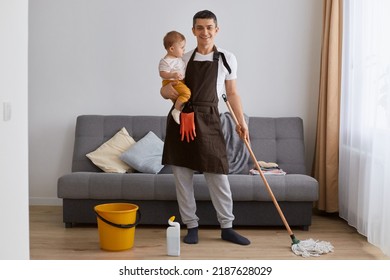 Horizontal Shot Of Smiling Delighted Attractive Man In Brown Apron Holding Baby Daughter In Hands And Washing Floor With Mop In Living Room, Looking At Camera.