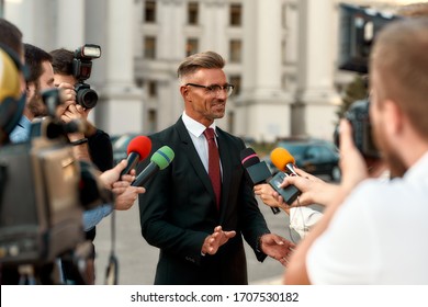 Horizontal Shot Of Smiling Politician Looking At Reporters And Giving Interview To Them. People Making Interview Using Microphones And Equipment Set At Outdoor Location. Selective Focus. Front View