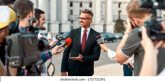 Horizontal Shot Of Smiling Politician Looking At Reporters And Giving Interview To Them. People Making Interview Using Microphones And Equipment Set At Outdoor Location. Selective Focus. Front View