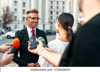 Horizontal Shot Of Smiling Politician Looking At Female Reporter And Giving Interview To Her. People Making Interview Using Microphones And Equipment Set At Outdoor Location. Selective Focus