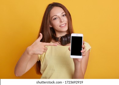 Horizontal shot of smiling woman pointing at blank device's screen holding in hands, lady with headphones around neck wearing casual t shirt, girl with long dark hair, against yellow studio wall.