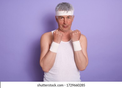 Horizontal Shot Of Strong Confident Senior Man Wearing White Sport Shirt And Headband, Keeping Hands In Fists, Boxing, Learning Kick, Being Focused, Having Determined Facial Expression. Sport Concept.