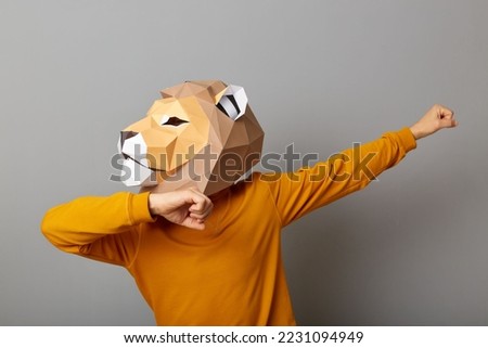 Horizontal shot of strong man with lion mask with wearing casual style orange jumper posing isolated over gray background, standing with raised arms and clenched fists, looking away.
