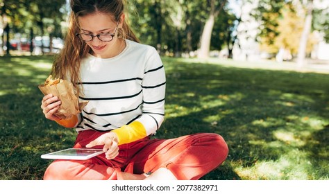 Horizontal Shot Of A Student Female In Casual Outfit, Eyeglasses, Sitting On The Green Grass At The College Campus, Have Lunch, And Studying. Woman Takes A Rest Eating Fast Food And Learning Outside.