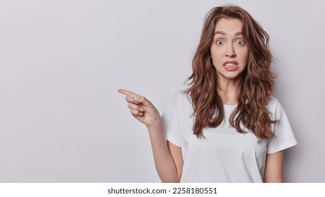 Horizontal shot of stunned embarrassed woman with long wavy hair stares at camera clenches teeth points aside on blank space demonstrated something awesome dressed casually isolated over white wall