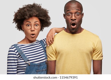 Horizontal shot of stunned emotive dark skinned family couple doesn`t expect to recieve bills, keep jaw dropped, see no way out, have no money to pay, isolated over white background. Omg concept