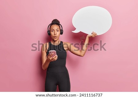 Horizontal shot of stunned Latin sporty woman dressed in black sportswear holds smartphone and speech bubble for your advertising content stares wondered at camera isolated over pink background