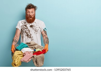 Horizontal Shot Of Stupefied Housekeeper Carries Laundry Basket, Has Dirty Clothing, Tattooed Arms, Shocked To Have Much Washing, Stands Against Blue Wall With Empty Space For Your Slogan Or Text