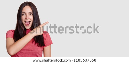 Horizontal shot of surprised brunette woman with dark hair, dressed in pink t shirt, points with index finger asie, shows free space for your promotion, notices something unbelievable. Look there!