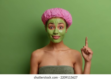 Horizontal Shot Of Surprised Young Female Model Points Index Finger Above Wears Waterproof Bathhat And Towel Around Body Applies Nourishing Clay Mask To Reduce Fine Lines Poses Against Green Wall