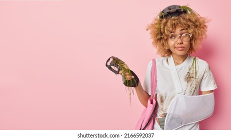 Horizontal Shot Of Thoughtful Curly Haired Young Woman Has Broken Arm After Dangerous Riding On Bicycle Stops On Road Holds Pedal Has Breakdown Of Transport Wears Protective Gear Poses Indoor