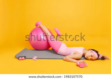 Horizontal shot of tired exhausted woman lies on mat keeps les on fitness ball feels very tired after workout trains at home dressed in sportive outfit exercises in room isolated on yellow background