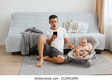 Horizontal Shot Of Tired Sleepy Man Wearing White Casual Style T-shirt And Jeans Short Sitting On Floor With Baby In Rocking Chair, Yawning And Using Cell Phone.