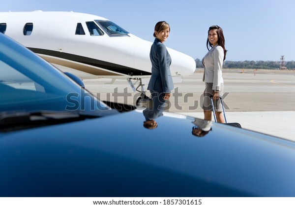 Horizontal shot of two businesswomen by car and\
airplane on a runway at the\
airport.
