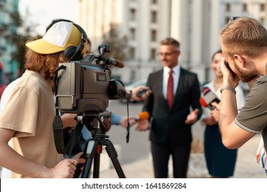 Horizontal Shot Of Two Operators Recording And Shooting Interview Of Politician. People Making Interview Using Photo- And Video- Cameras Set At Outdoor Location. Selective Focus On Cameraman