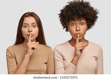 Horizontal Shot Of Two Shocked Women Of Different Nationalities Make Silence Gesture, Gossip About Something Very Private, Stand Closely, Isolated Over White Background. People And Diversity Concept