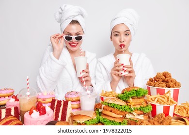 Horizontal Shot Of Two Women Drink Fizzy Drink With Straws Wear Domestic Comfortable Dressing Gown Cant Resist Craving For Junk Food Pose Near Table Full Of High Calorie Cakes Burgers French Fries