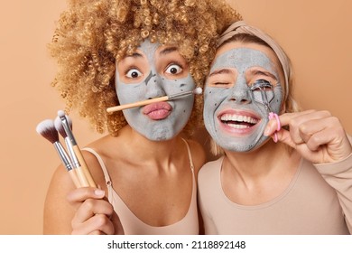 Horizontal shot of two women undergo beauty procedures apply clay mask use cosmetic brushes and eyelashes curler isolated over brown background prepare for party. Skin care cosmetology concept