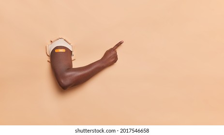 Horizontal shot of unknown dark skinned person breaks arm through beige paper background points away in copy space shows place for your advertising content makes vaccination shot in shoulder
