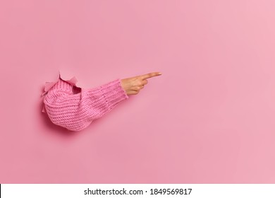 Horizontal shot of unrecognizable woman points away on copy space breaks through pink paper studio wall dressed in sweater. Shopping proposal advertising and discounts concept. Break throughout