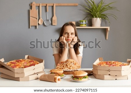 Horizontal shot of upset bored Caucasian woman wearing white casual T-shirt sitting at table in kitchen, keeps hand under chin, looking at junk food, can't eat, keeps diet.