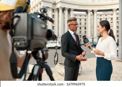 Horizontal Shot Of Video Camera Operator Recording Interview Of Politician. People Making Interview Using Equipment Set At Outdoor Location. Selective Focus