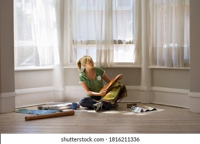 Horizontal Shot Of A Woman Decorating Home Sitting On Floor Beside Window And Holding Wallpaper.