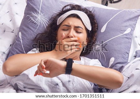 Horizontal shot of yawning girl lying in bed on pillow, under blanket, looking at her smart watch and covering her mouth with hand, wakes up early in morning, does not need go anywhere, staying home.