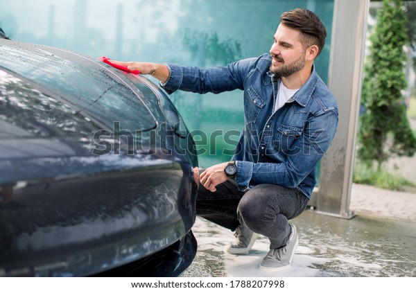 Horizontal shot of a young\
bearded man in jeans shirt, cleaning the hood on his luxury modern\
electric car outdoor with red microfiber cloth, at self service car\
wash