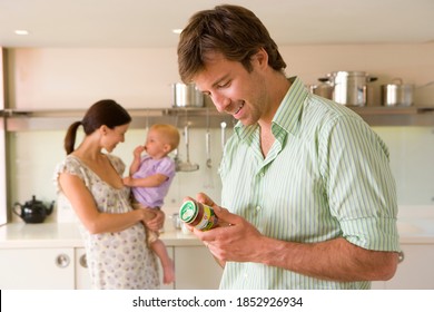 Horizontal Shot Of A Young Couple In Kitchen With The Pregnant Woman Holding A Baby Girl And Man In The Foreground Reading Label On A Baby Food Bottle.