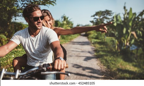 Horizontal Shot Of Young Couple Riding Motorbike. Man Riding On A Motorcycle With Girlfriend Pointing Away On Rural Road.