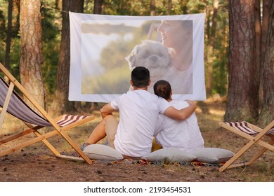 Horizontal Shot Of Young Loving Couple Sitting In The Forest With Overhead Projector, Hugging Each Other And Looking At Projector Screen, Spending Time In Open Air In Beautiful Nature.