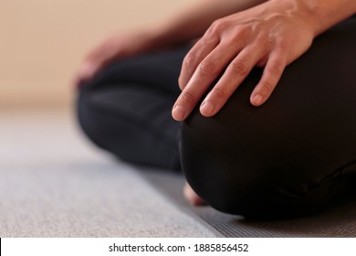Horizontal side closeup of lower body of yogini on the floor with legs crossed in lotus pose. Woman indoors wearing black yoga pants with hands resting on knees. Home yoga practice