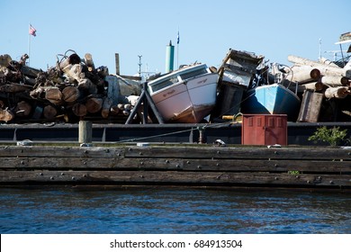 Horizontal side perspective of collected garbage of dead tree logs, storm debris, and abandoned vintage beached motor boats piled on wood dock at harbor above water in industrial waterway port scene - Powered by Shutterstock