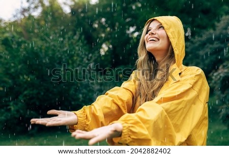 Horizontal side view image of a joyful young woman smiling wearing yellow raincoat during the rain in the nature. Pretty female looking up and catching the rain drop with hands outdoors in the park.