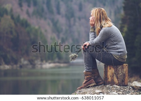 horizontal side view portrait of Caucasian young blonde woman with light colored sweater and jeans holding tree branch and meditating relaxing alone on tree stump front of lake surrounded by forest 