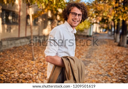 Horizontal side view potrait of young businessman with glasses posing outdoors. College male student in autumn street. Smart guy in casual wears spectacles with curly hair walking down the street